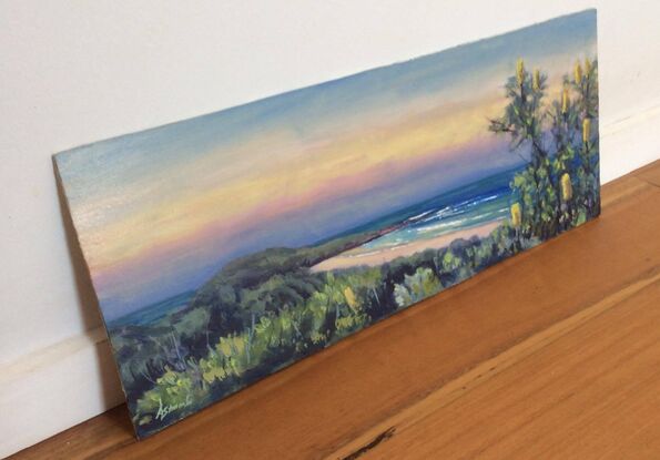 View towards Lover's Point, Pippi Beach, Yamba, at sunset. The sky is filled with soft yet vibrant colours like pink, yellow and blue, and to the right is a Banksia tree with yellow blossoms. Gentle waves curl into shore.