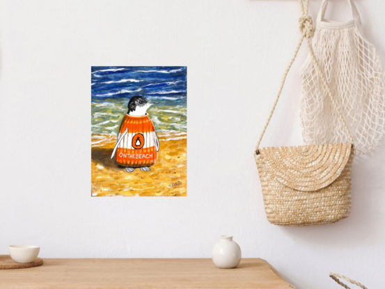 A solitary penguin, wearing a knitted orange and white jumper resembling a Penguin Classic book, features the title "On The Beach" inspired by Neville Shute's novel. This lone penguin stands at the shoreline, perfectly embodying the playful wordplay of being "on the beach."