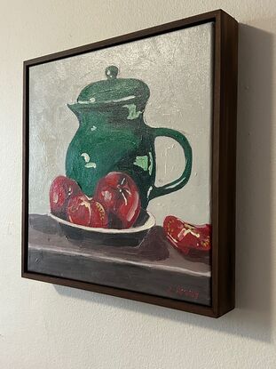 A emerald green jug sits on a timber bench and in the foregground is a bowl with three tomatoes. The background is a off white