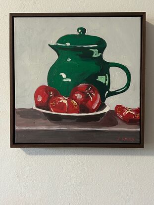 A emerald green jug sits on a timber bench and in the foregground is a bowl with three tomatoes. The background is a off white