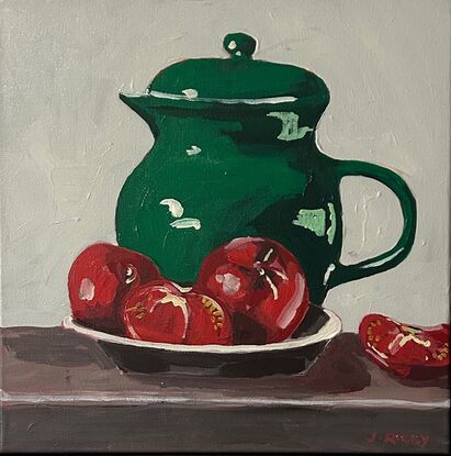 A emerald green jug sits on a timber bench and in the foregground is a bowl with three tomatoes. The background is a off white