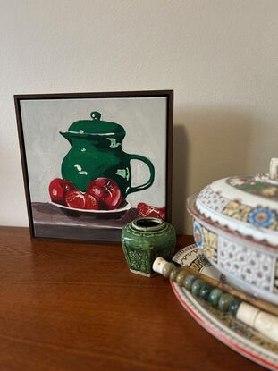 A emerald green jug sits on a timber bench and in the foregground is a bowl with three tomatoes. The background is a off white