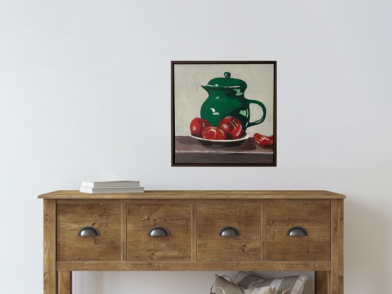 A emerald green jug sits on a timber bench and in the foregground is a bowl with three tomatoes. The background is a off white