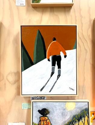 A lone male skier is view from behind. He is wearing an orange jackets and black pants and ski boots. He is trekking up the white snow hill towards some green conifers in the distance. The sky above is a warm orangey brown colour. 