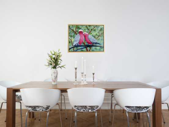 Two pink and grey galahs sitting on a branch in a loving and romantic embrace. One galah appears to be grooming its mate. The background is various shades of green representing the leaves in the trees behind them and blue sky filters through the leaves. 