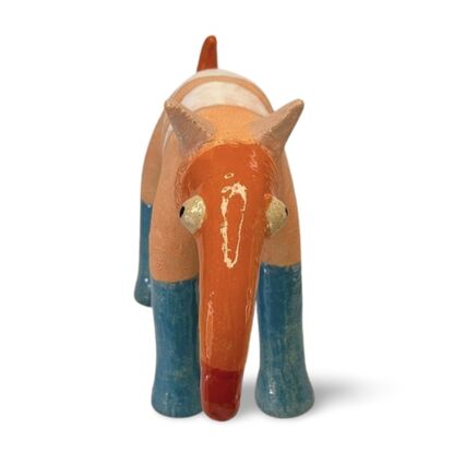 The Peachy Snoutosaurus is a charming ceramic creature with a rotund, striped body in soft orange hues, balanced on four sturdy blue legs. Its most striking feature is an elongated, trunk-like snout that curves dramatically upward, ending in a vibrant red tip. The eyes give this whimsical sculpture a watchful and endearing presence, enhancing its playful and curious character.