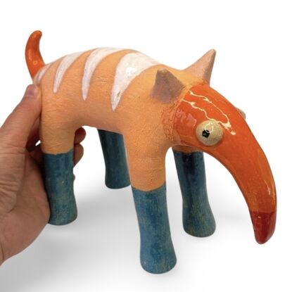 The Peachy Snoutosaurus is a charming ceramic creature with a rotund, striped body in soft orange hues, balanced on four sturdy blue legs. Its most striking feature is an elongated, trunk-like snout that curves dramatically upward, ending in a vibrant red tip. The eyes give this whimsical sculpture a watchful and endearing presence, enhancing its playful and curious character.