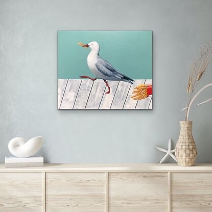 A seagull with chips in its beak marching away from a paper cup of fries laying on a whiter wooden table.