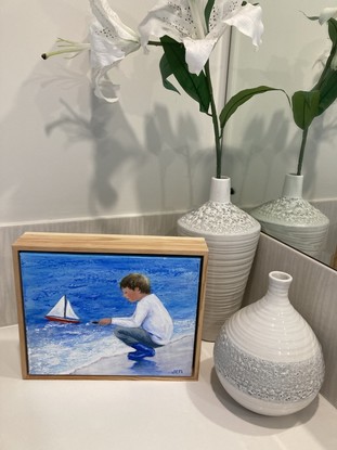 Young boy sailing his new boat for the first time. Main colours are blue and white with a touch of mauve and pink in the sand where his blue boots are reflected.