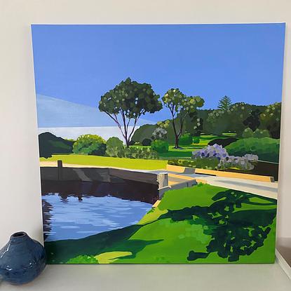 Shadows on grass beside blue lake with bridge and trees in background with blue sky